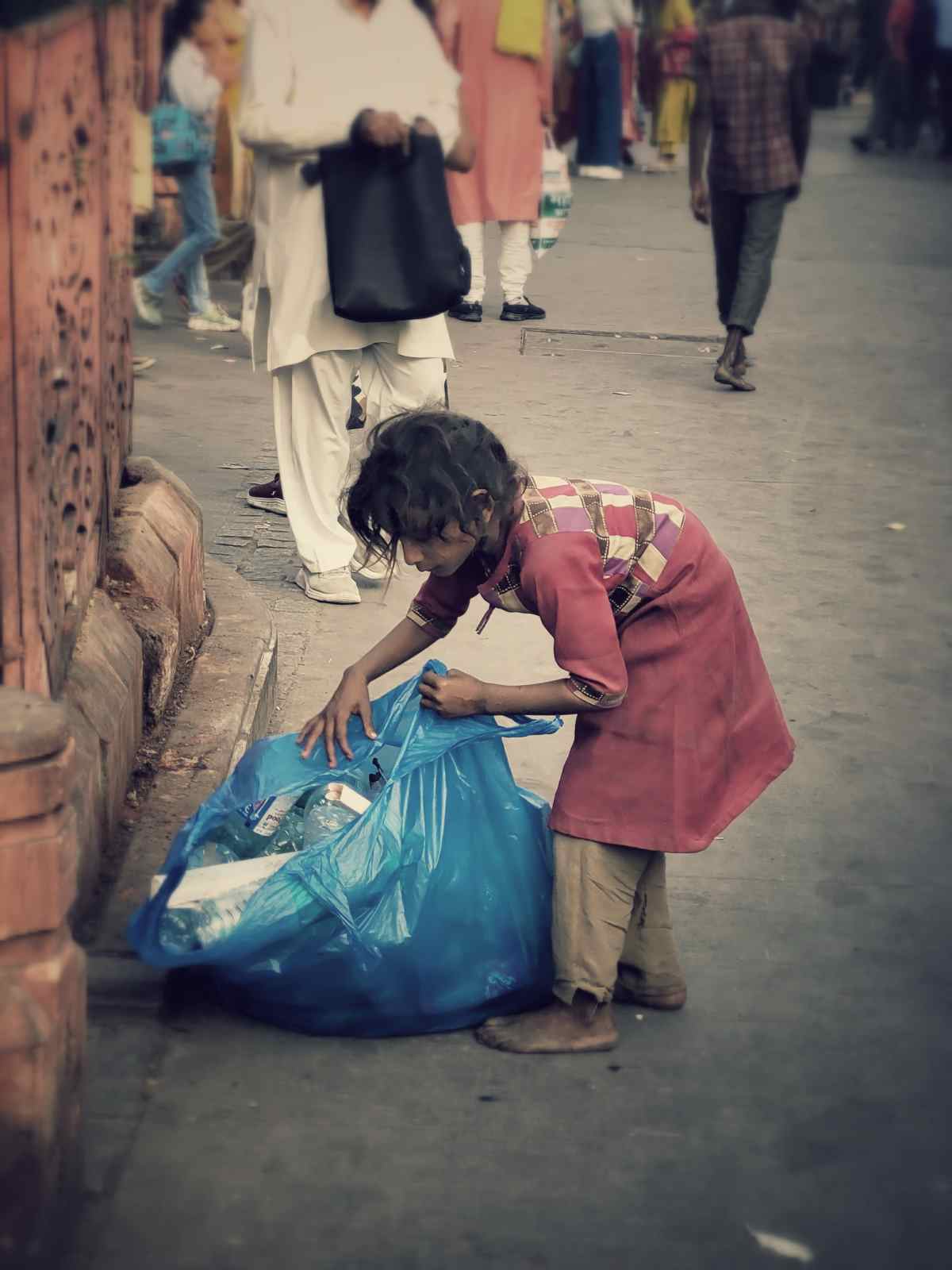 Children: Scraping Together a Living: A little girl combs through the streets, gathering waste to earn a few rupees. Her small hands work tirelessly, each piece collected a step toward supporting her family’s needs