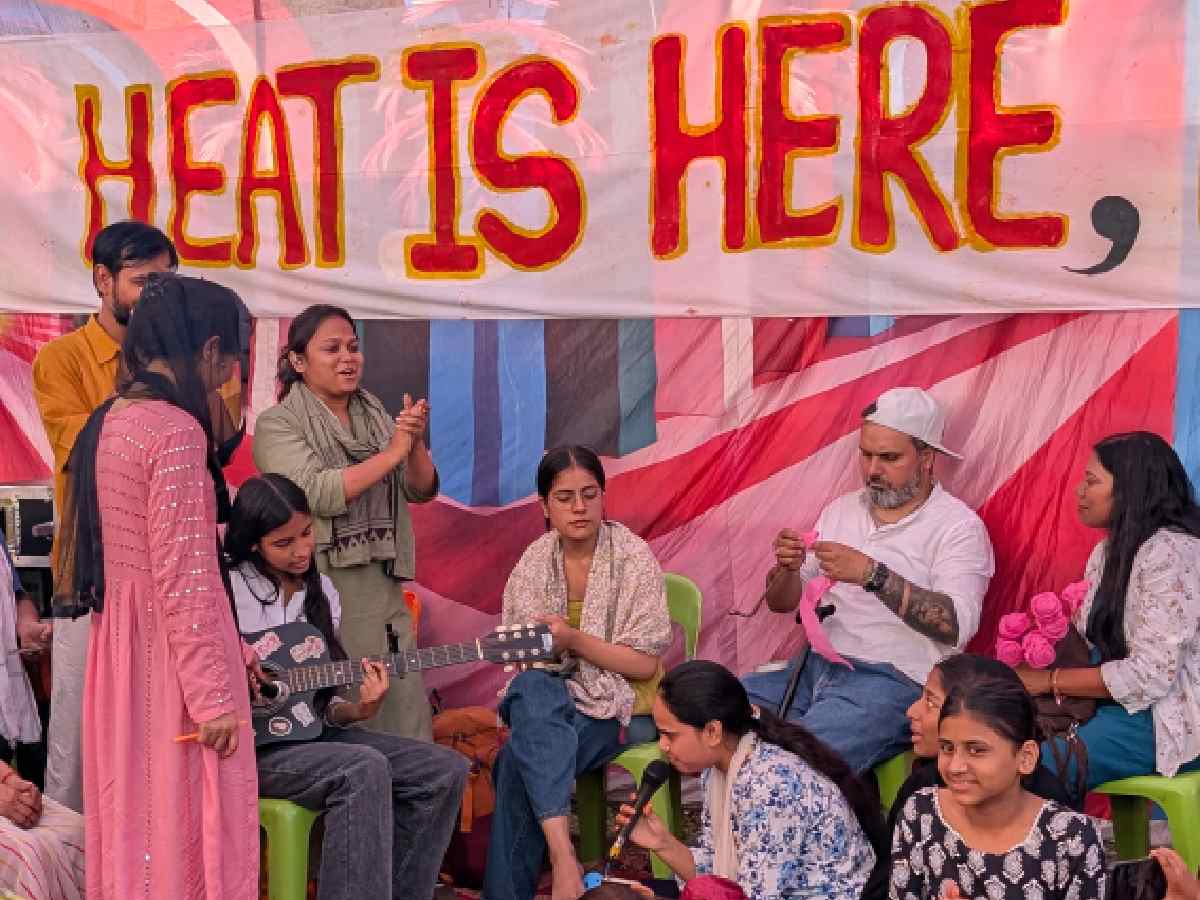 A group of young performers sing and play music during a gathering, as a backdrop reading “Heat is Here” highlights concerns